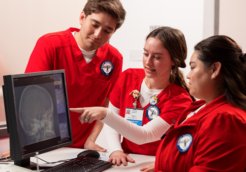 Three radiologic tech students (1 male and 2 females) in red scrubs look at a digital x-ray. 