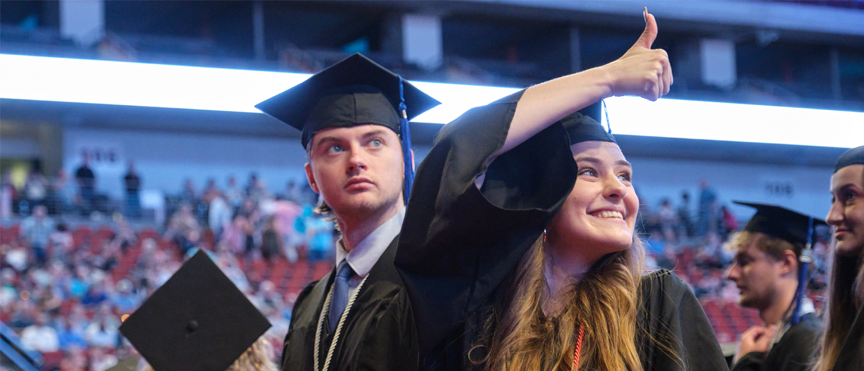 Female graduate with long blonde hair gives a thumbs up as a male graduate looks on. 