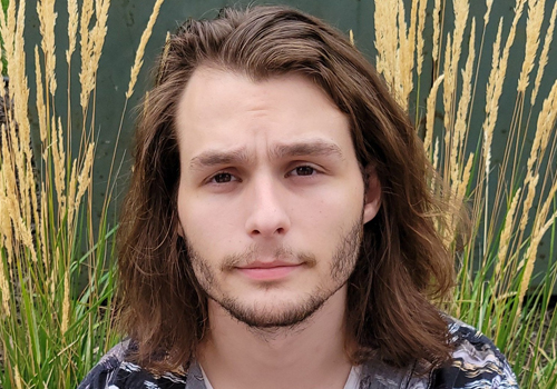 Levi Chandler in an outdoor setting. He has shoulder length, brown hair, brown eyes and beard. 